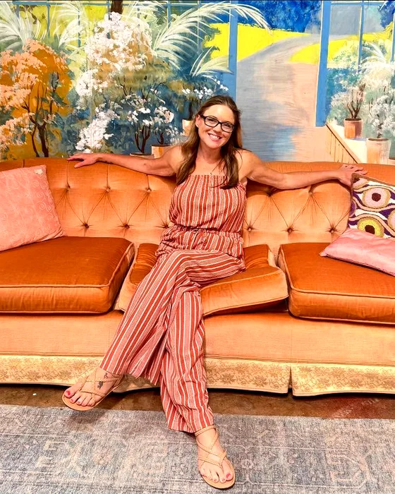 Lindsay Knowles, owner of LNK Creative, smiling on an orange velvet sofa in front of a painted garden mural