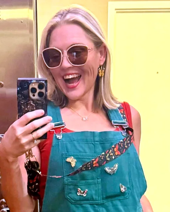 Lindsay Knowles, owner of LNK Creative, laughing in an elevator mirror selfie, wearing green overalls with butterfly pins